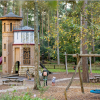 Woodland playground with wooden castle tower, slides, swings and picnic tables, surrounded by tall trees and children playing