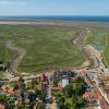 Aerial view of a coastal village with winding river and lush green marshland leading to the open sea in the distance