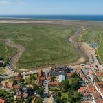 Aerial view of a coastal village with winding river and lush green marshland leading to the open sea in the distance
