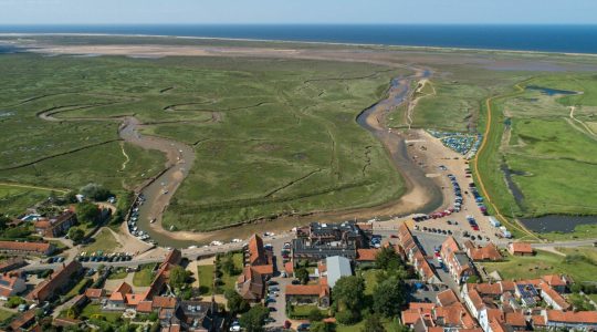 Aerial view of a coastal village with winding river and lush green marshland leading to the open sea in the distance