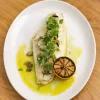 Grilled fish fillet with microgreens and charred lemon on a plate, garnished with olive oil, on a wooden table