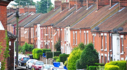 Street view of traditional red brick terraced houses with parked cars and green hedges in a British neighbourhood