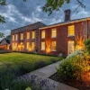 Historic brick house with warm evening lights, lush garden, and cloudy sky in a tranquil setting