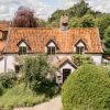 Charming countryside cottage with orange roof surrounded by lush green trees and shrubs under a cloudy sky