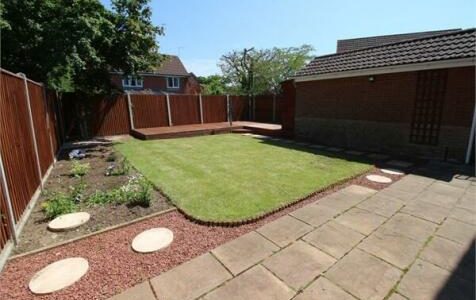 Back garden with green lawn, paved patio, and wooden deck surrounded by red fences and a small flowerbed