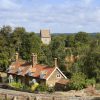 Charming English countryside scene with red-roofed cottage, lush green trees, and distant church tower on a sunny day