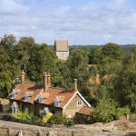 Charming English countryside scene with red-roofed cottage, lush green trees, and distant church tower on a sunny day
