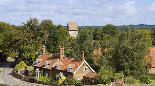 Charming English countryside scene with red-roofed cottage, lush green trees, and distant church tower on a sunny day