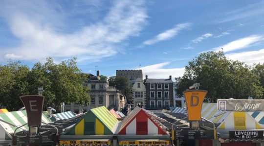 Colourful market stalls with vintage clothing signage under a clear blue sky in a historic city setting