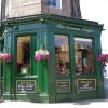 Green corner pub called The Conan Doyle with hanging flowers and clear windows displaying the interior, sunlight reflecting