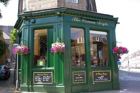 Green corner pub called The Conan Doyle with hanging flowers and clear windows displaying the interior, sunlight reflecting
