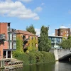 Modern riverside buildings in a picturesque setting with a stone bridge and lush greenery under a bright blue sky