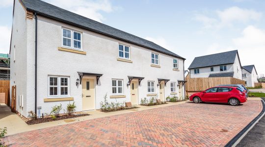 Newly built semi-detached homes with red car parked on brick driveway under clear blue sky in suburban UK neighbourhood