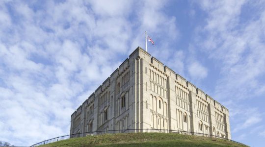 Norwich Castle under a clear blue sky with a flag flying on top, set on a grassy hill with a surrounding fence