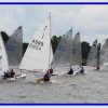 Sailing boats racing on a lake with sailors in action, surrounded by trees under a cloudy sky in a vibrant scene