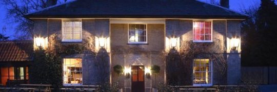 Historic English inn lit up at night, showcasing warm lighting and classic architecture, surrounded by benches and greenery