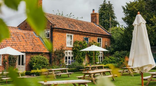 Cosy English pub garden with red brick house, picnic tables, and parasol amidst lush greenery