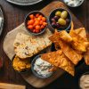 Assortment of breads, olives, dips, and chips on a wooden board with a refreshing drink and the edge of a frothy beer
