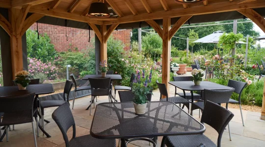 Outdoor patio with tables and chairs under a wooden pergola surrounded by lush garden flowers and greenery