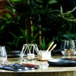 Elegant restaurant table setting with empty glasses, cutlery, candleholder, and plants in the background