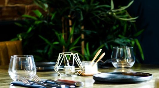 Elegant restaurant table setting with empty glasses, cutlery, candleholder, and plants in the background