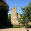 Medieval brick tower with a yellow flag, surrounded by lush greenery and flowers on a sunny day in a historical setting