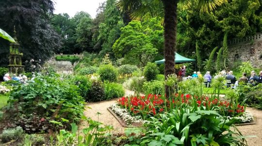 Lush English garden with vibrant flowers and people sitting under a green canopy surrounded by trees and greenery
