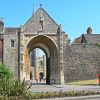 Historic stone gate entrance to Norwich Cathedral under a clear blue sky with surrounding greenery and vibrant flowers