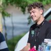 Man smiling outdoors with brochures and speaking to person in casual setting, wearing a name badge and lanyard