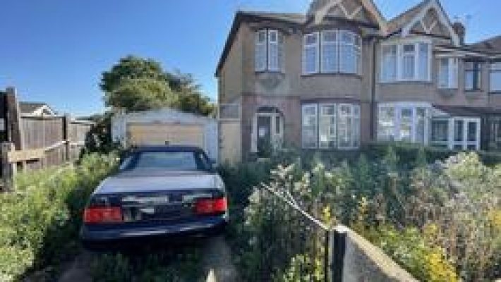 Overgrown garden with parked car beside a semi-detached house under a clear blue sky in a residential neighbourhood