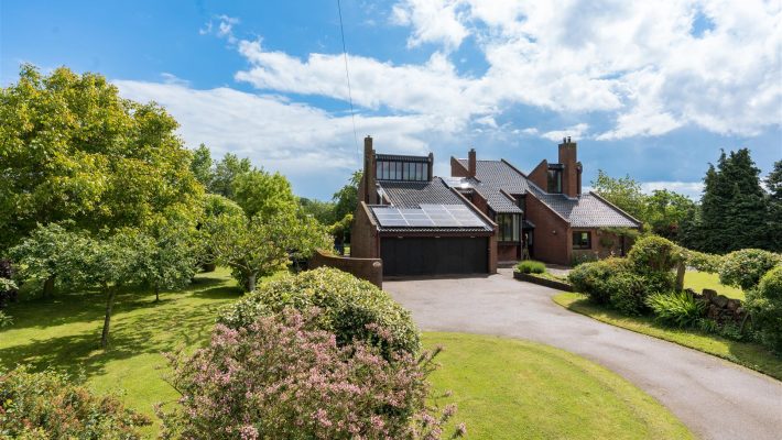 Modern brick house with solar panels surrounded by lush garden and trees under a bright blue sky in the UK countryside