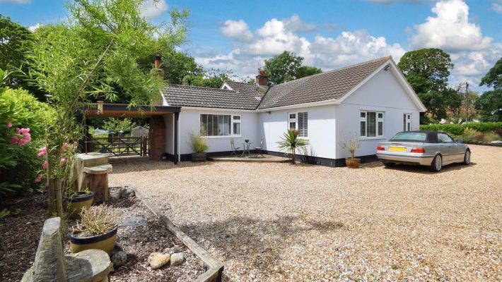 Charming white bungalow with gravel driveway, lush greenery, and a silver car in a sunny countryside setting