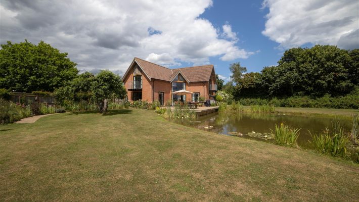 Brick house by a tranquil pond with lush garden under a partly cloudy sky in a countryside setting