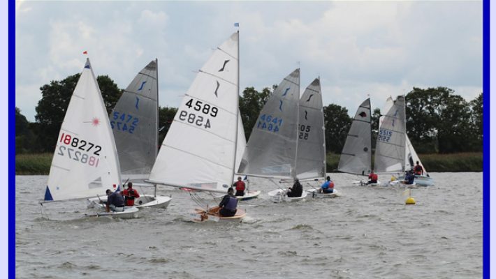 Sailing boats racing on a lake with sailors in action, surrounded by trees under a cloudy sky in a vibrant scene
