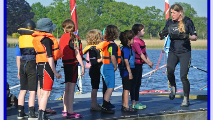 Instructor guiding children in colourful life jackets by a lake for a sailing lesson