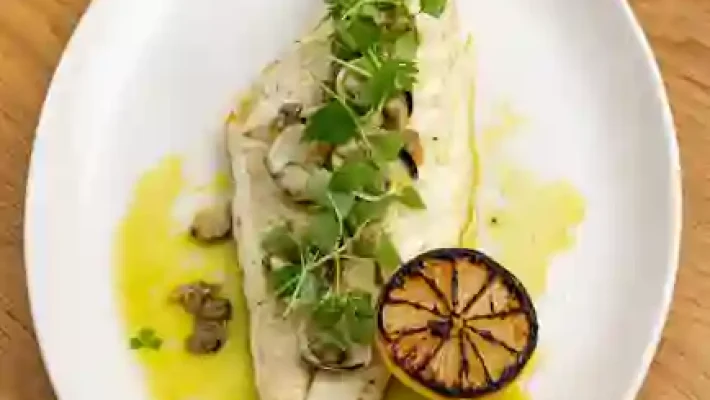 Grilled fish fillet with microgreens and charred lemon on a plate, garnished with olive oil, on a wooden table
