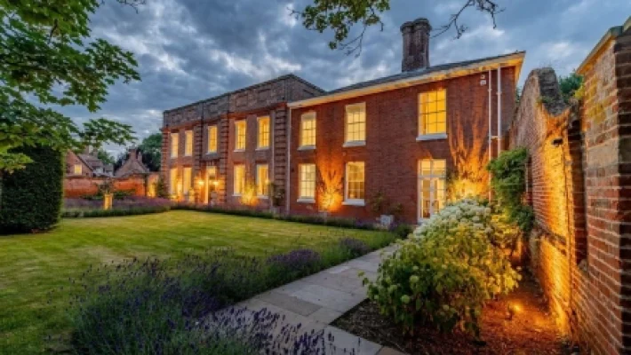 Historic brick house with warm evening lights, lush garden, and cloudy sky in a tranquil setting