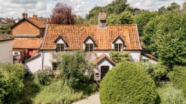 Charming countryside cottage with orange roof surrounded by lush green trees and shrubs under a cloudy sky