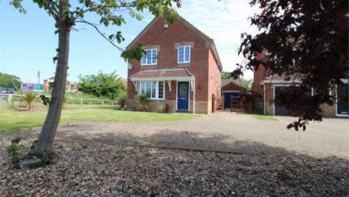 Brick house with blue door, gravel driveway, and tree in front garden on a sunny day in a suburban area