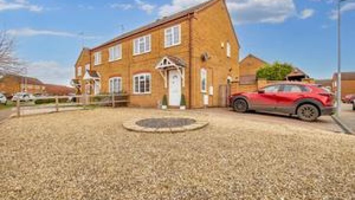 Red car parked beside a semi-detached house on a gravel driveway under a blue sky in a suburban neighbourhood