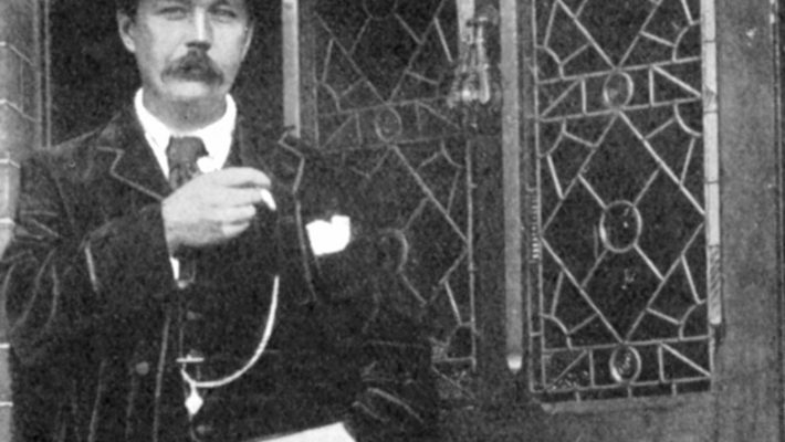 Man in vintage attire stands near ornate door, holding a pipe and wearing a hat, evoking early 20th-century style