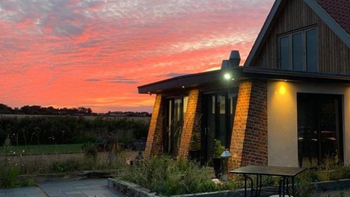 Modern house with red brick and wooden accents, set against a vibrant sunset sky in a peaceful rural landscape