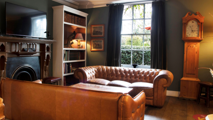 Cosy vintage living room with leather sofas, bookshelf, fireplace, grandfather clock, and large window view of greenery