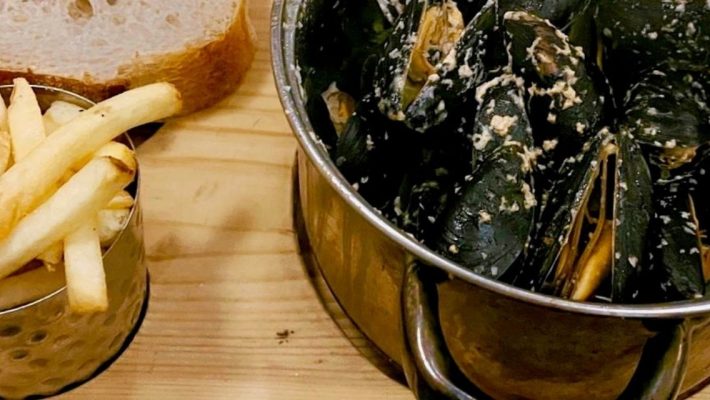 Mussels in a metal pot with a side of crispy chips and a slice of fresh bread on a wooden board
