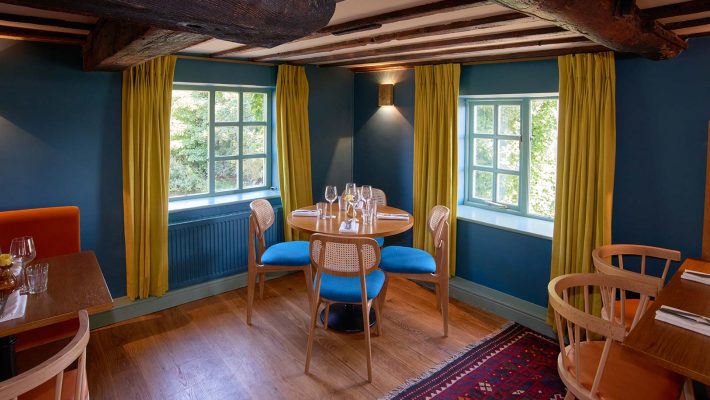 Cosy dining room with wooden furniture, blue chairs, yellow curtains and exposed beams, natural light through windows