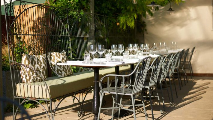 Outdoor dining area with elegant table setting, green bench, and black metal chairs under lush foliage