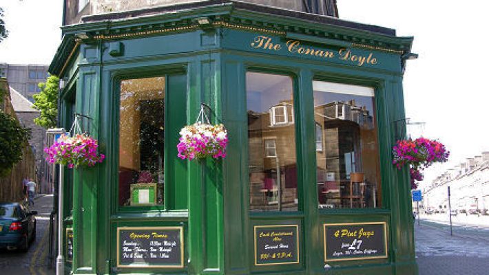 Green corner pub called The Conan Doyle with hanging flowers and clear windows displaying the interior, sunlight reflecting
