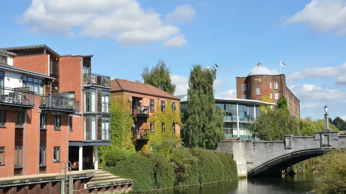 Modern riverside buildings in a picturesque setting with a stone bridge and lush greenery under a bright blue sky