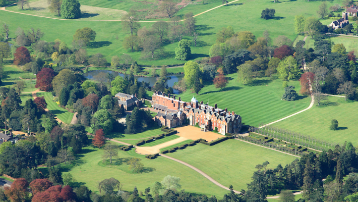 Aerial view of a historic English manor house surrounded by lush green gardens and trees in springtime beauty