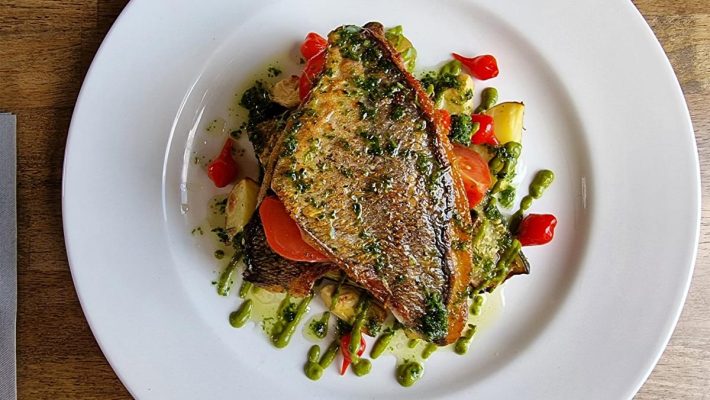 Grilled fish on a plate with sliced tomatoes, garlic, and pesto, served on wooden table background